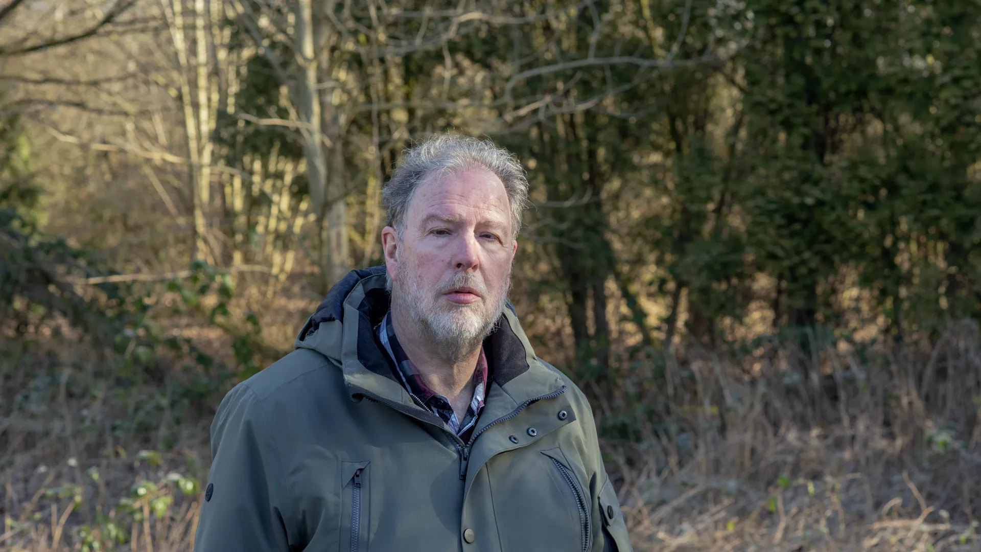 Man in jas staat buiten in een bosrijke omgeving.