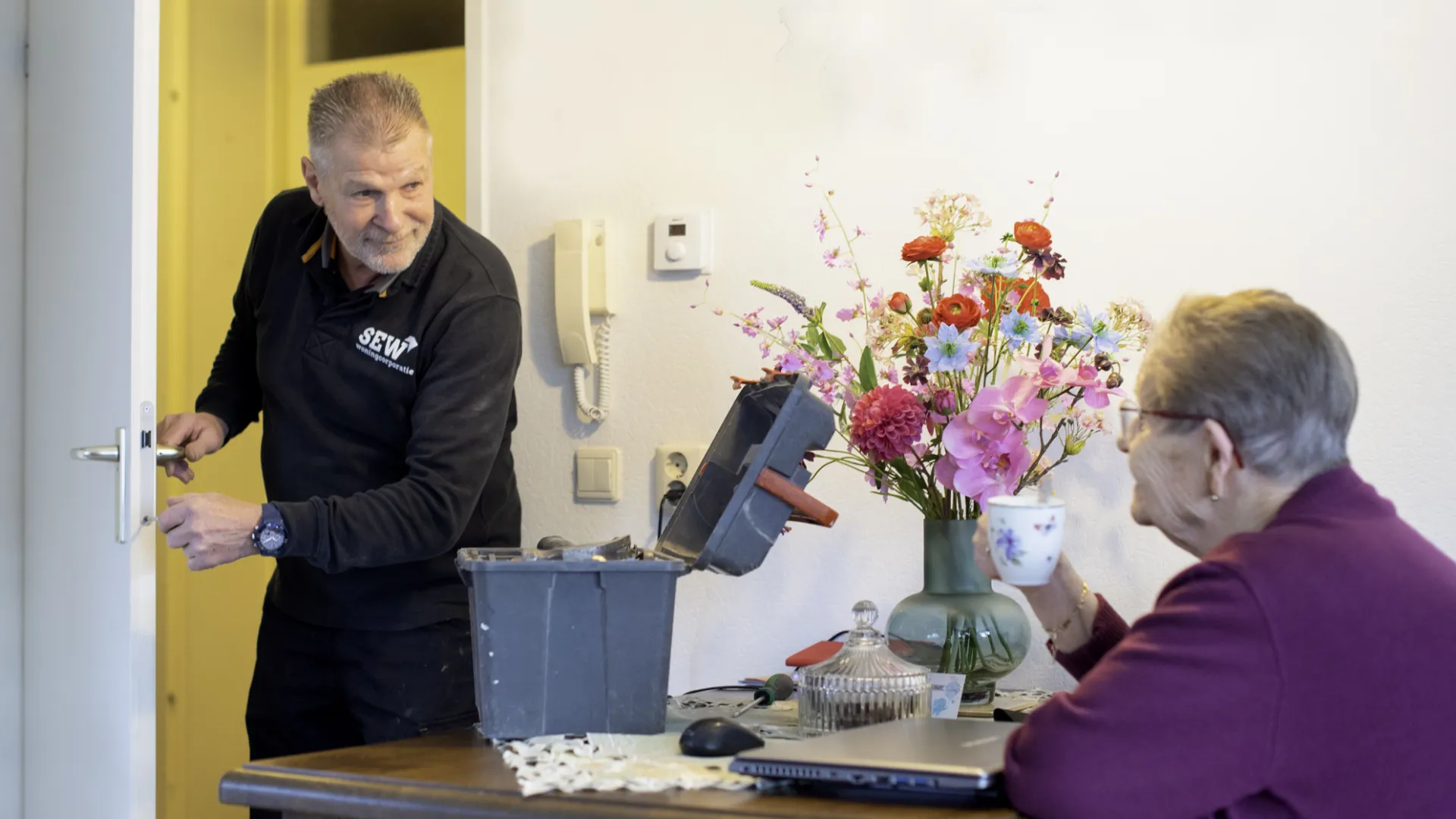Monteur van Stichting Eelder Woningbouw repareert een deur terwijl een huurder toekijkt.