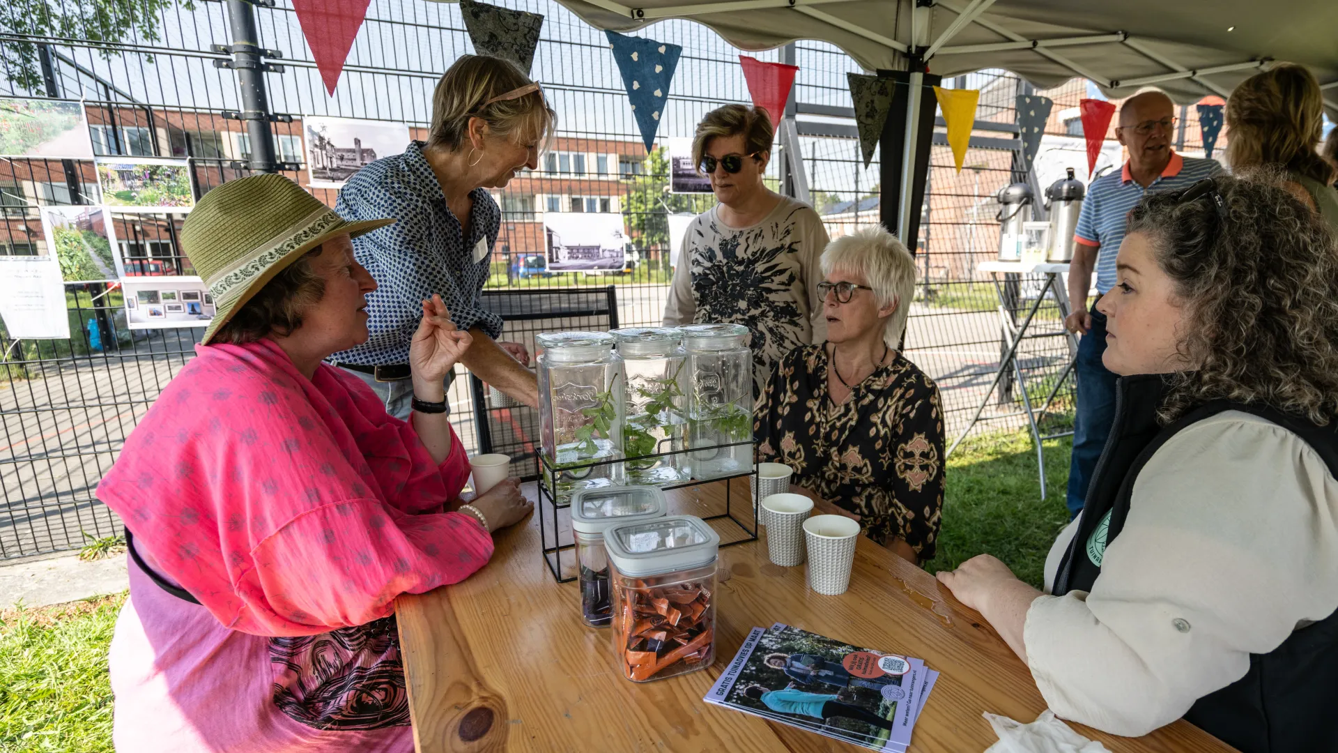 Mensen zitten en praten aan een tafel tijdens een buitenactiviteit met een kraampje en vlaggetjes.