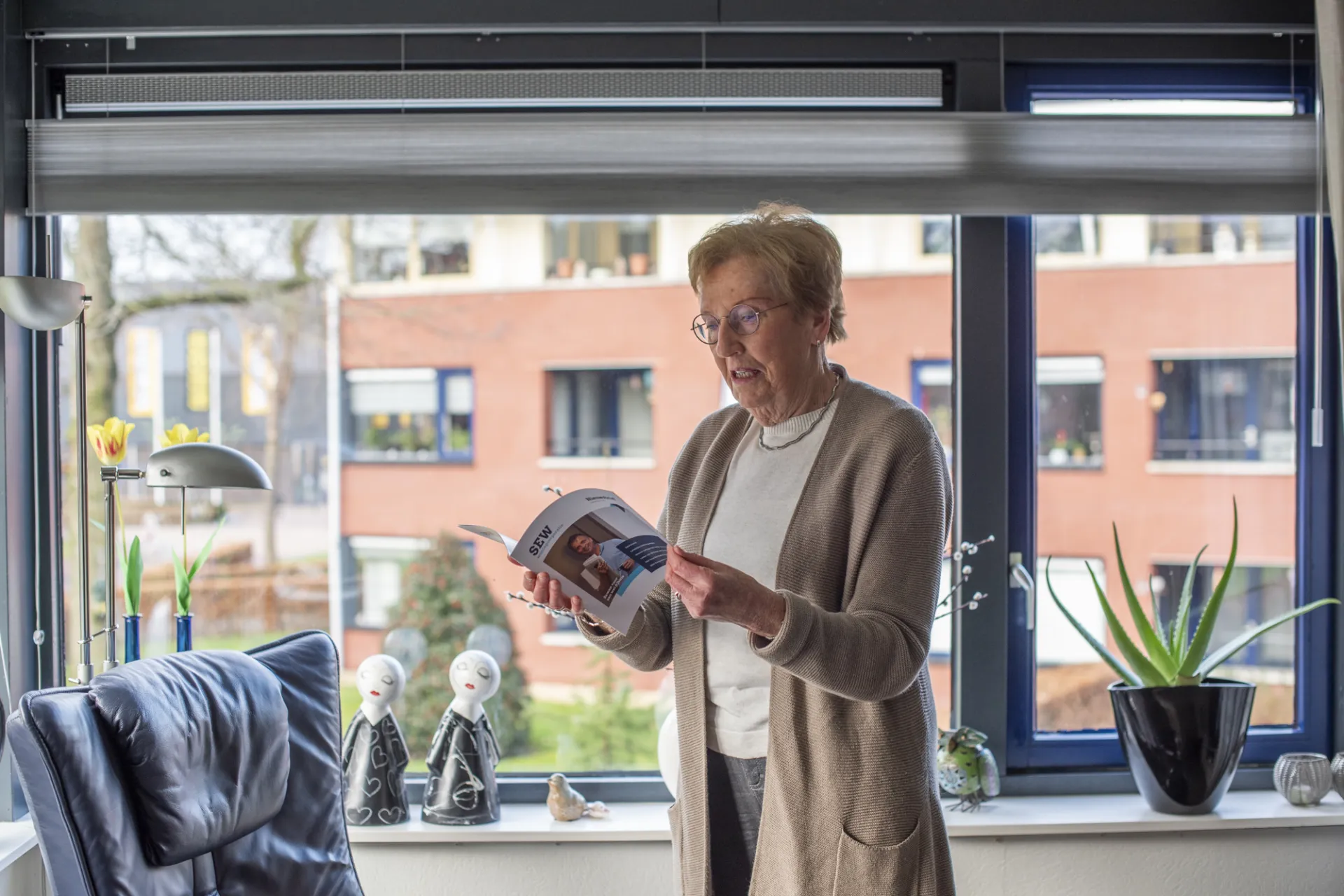 Een oudere vrouw leest een boek bij het raam in een gezellig ingerichte kamer.