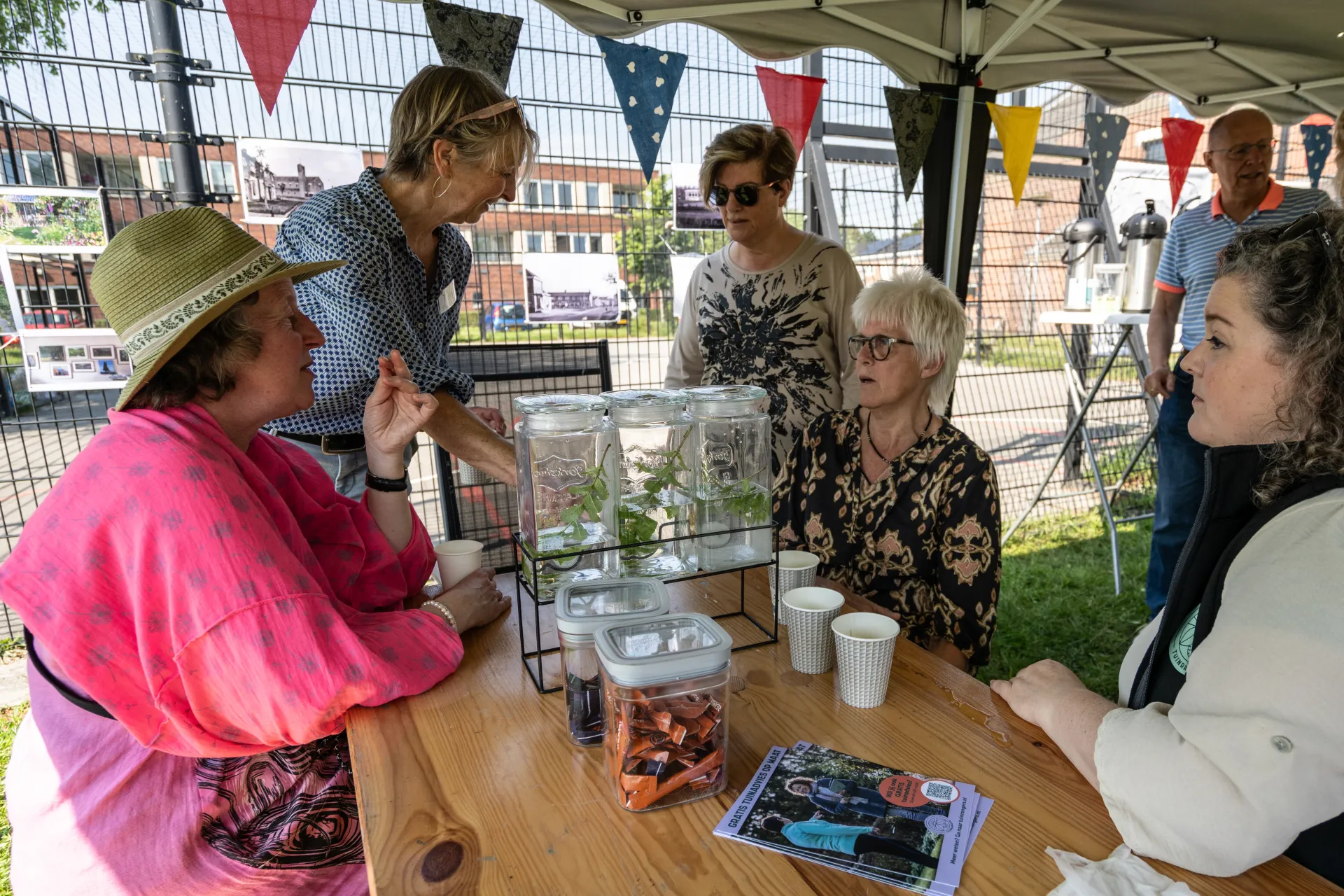 Mensen zitten en praten aan een tafel tijdens een buitenactiviteit met een kraampje en vlaggetjes.